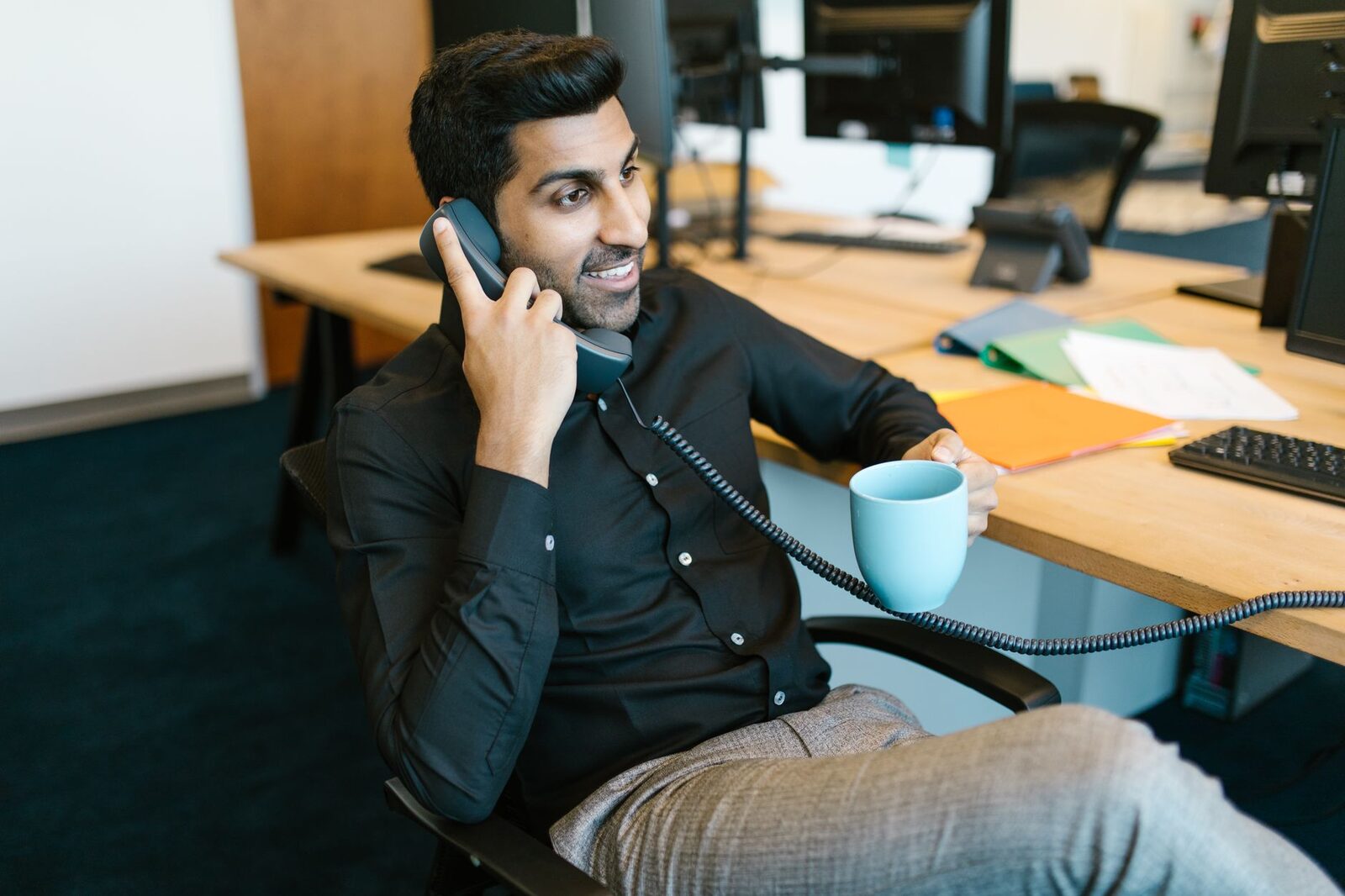Smiling man sits in office holding a mug and talking on the phone, conveying a professional environment.